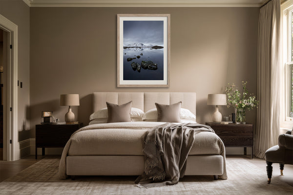 Light wood-framed mountain lake artwork centered above a beige upholstered bed.