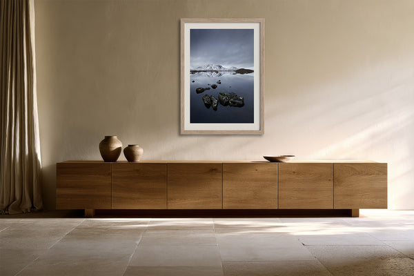Light wood-framed mountain lake artwork above a low wooden sideboard with ceramic vessels.