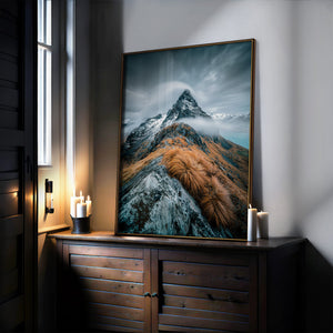 Large mountain artwork in a thin dark frame leaning on a wooden cabinet beside candles.