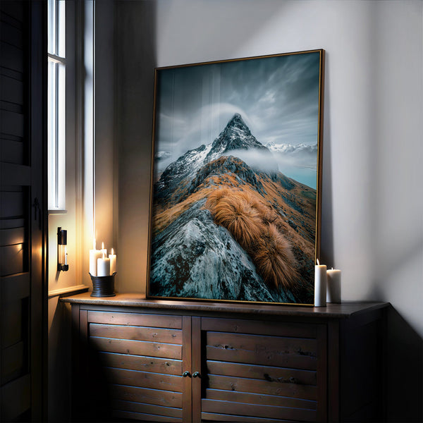 Large mountain artwork in a thin dark frame leaning on a wooden cabinet beside candles.