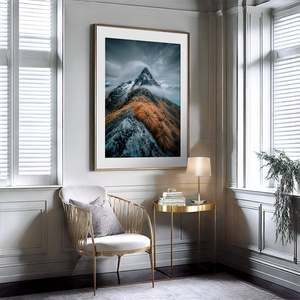 White-matted mountain print in a narrow gold frame above a chair and side table.