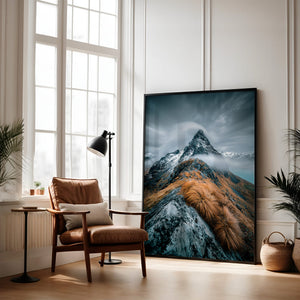 Large dark-framed mountain artwork leaning beside a brown armchair and floor lamp.