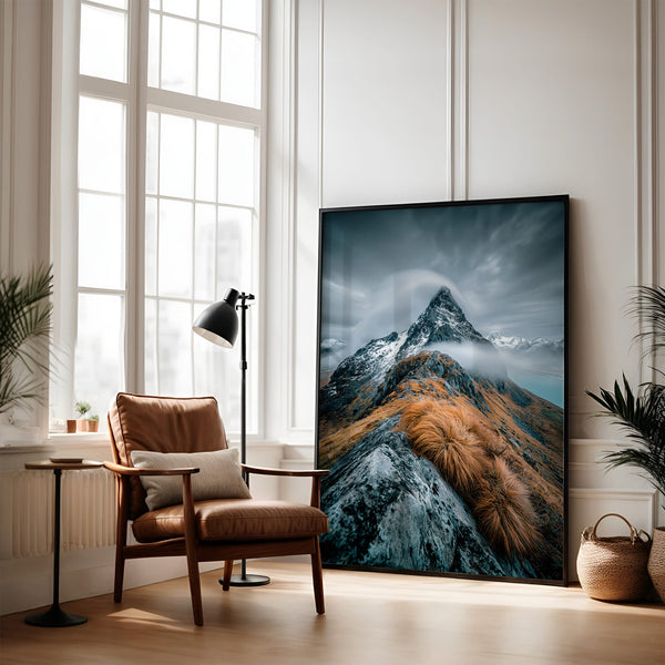 Large dark-framed mountain artwork leaning beside a brown armchair and floor lamp.
