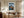 Framed mountain print above a wooden desk and office chair beside tall curtains.