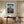 Framed mountain print above a wooden desk and office chair beside tall curtains.