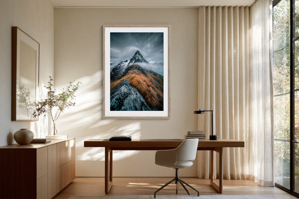 Framed mountain print above a wooden desk and office chair beside tall curtains.