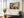 Framed geometric owl artwork above a cushioned wooden bench near tall windows and a vase of dried flowers.