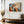 Framed geometric owl artwork above a cushioned wooden bench near tall windows and a vase of dried flowers.