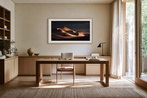 White-framed desert dunes artwork above a wooden desk beside tall windows and sheer curtains.
