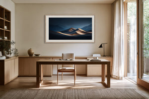 Light wood-framed desert dunes artwork above a wooden desk near tall glass doors.