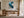 Framed blue and grey abstract artwork above a wooden desk near tall glass doors.
