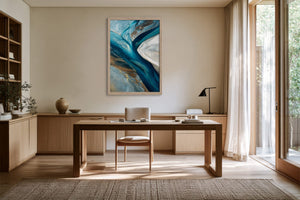 Framed blue and grey abstract artwork above a wooden desk near tall glass doors.