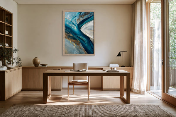 Framed blue and grey abstract artwork above a wooden desk near tall glass doors.