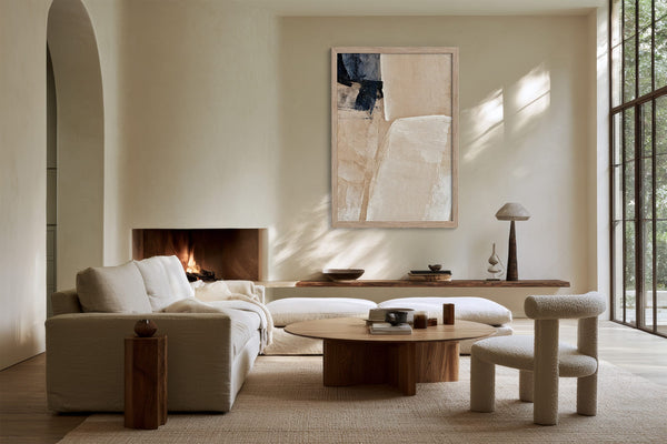 Framed beige and charcoal abstract artwork above a fireplace in a sunlit seating area.