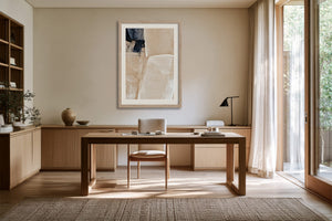 Framed beige and charcoal abstract artwork above a wooden desk beside tall windows.