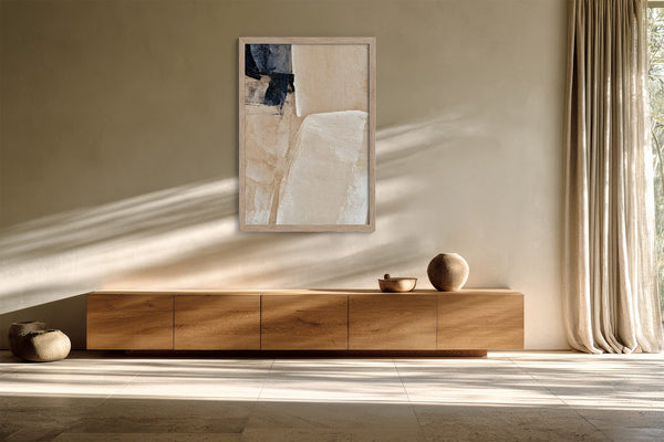 Framed beige and charcoal abstract artwork above a low wooden sideboard in filtered sunlight.