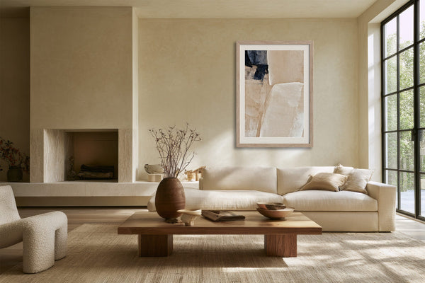 Framed beige and charcoal abstract artwork above a cream sofa near a fireplace and grid windows.