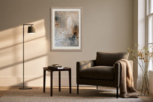 Framed beige, gray, and charcoal abstract artwork above an armchair beside a floor lamp.