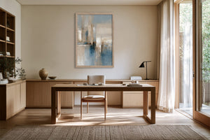 Framed blue and beige abstract artwork hung above a wooden desk beside tall windows.