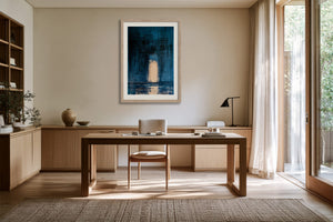 Framed blue abstract artwork with a pale arch displayed above a wooden desk.
