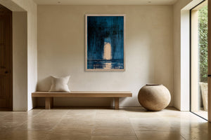 Framed blue abstract artwork with a pale arch displayed above a wooden bench and round vessel.