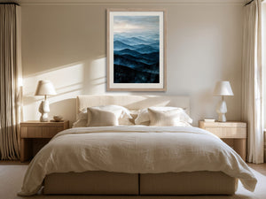 Light wood-framed blue mountain artwork centered above a bed between matching bedside lamps.
