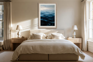 Light wood-framed blue mountain artwork centered above a bed between matching bedside lamps.