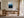 Light wood-framed blue mountain artwork displayed above a wooden desk in a sunlit workspace.