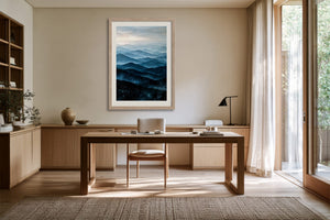 Light wood-framed blue mountain artwork displayed above a wooden desk in a sunlit workspace.