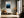Light wood-framed blue mountain artwork above a bench with an orange cushion near tall windows.