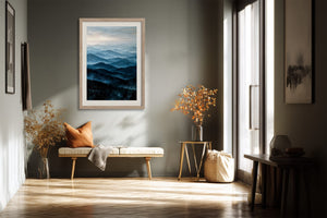 Light wood-framed blue mountain artwork above a bench with an orange cushion near tall windows.