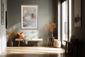 Framed pale abstract artwork above a bench with an orange cushion in a sunlit hallway.