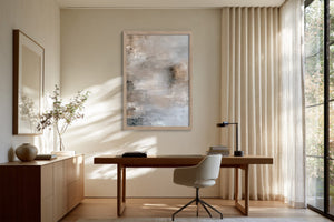 Framed gray-beige abstract artwork above a wooden desk in a bright workspace.