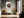Framed bird nest artwork with three speckled eggs above a cushioned bench near tall windows.