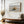 Dune grass coastal artwork in a thin wood frame above a cushioned bench and vase of dried flowers.
