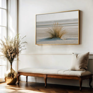 Dune grass coastal artwork in a thin wood frame above a cushioned bench and vase of dried flowers.