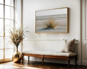 Dune grass coastal artwork in a thin wood frame above a cushioned bench and vase of dried flowers.