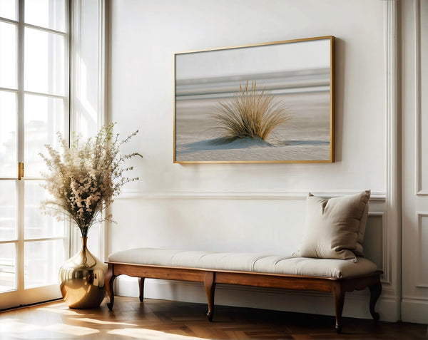 Dune grass coastal artwork in a thin wood frame above a cushioned bench and vase of dried flowers.