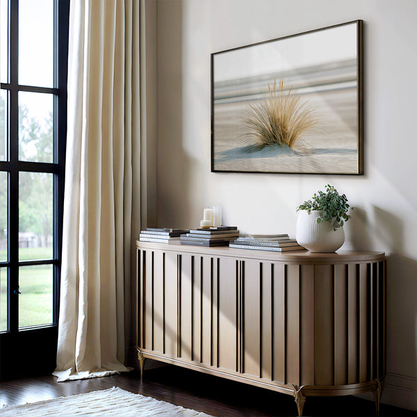 Framed dune grass artwork above a slatted console with stacked books, candles, and a potted plant.