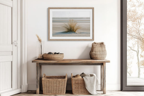 Dune grass artwork in a pale wood frame above a rustic console with woven baskets.