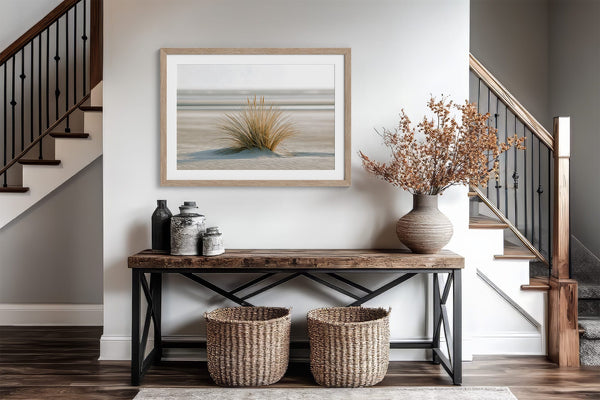 Dune grass artwork in a pale wood frame above a wood-and-metal console between staircases.