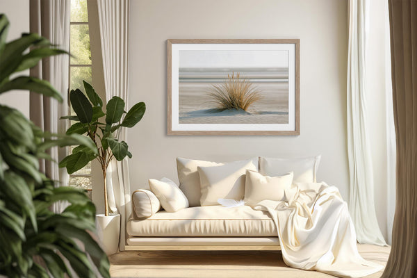 Dune grass artwork in a pale wood frame above a cream sofa with white cushions and curtains.