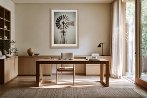 Framed windmill artwork above a wood desk and chair beside large windows.