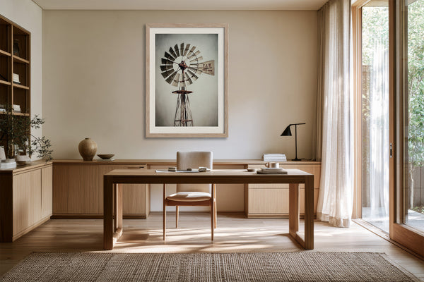 Framed windmill artwork above a wood desk and chair beside large windows.