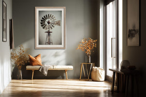 Light wood-framed windmill art above a bench with an orange pillow in a sunlit corridor.