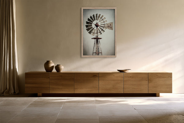 Framed windmill wall art above a long low wood cabinet with ceramic vessels.