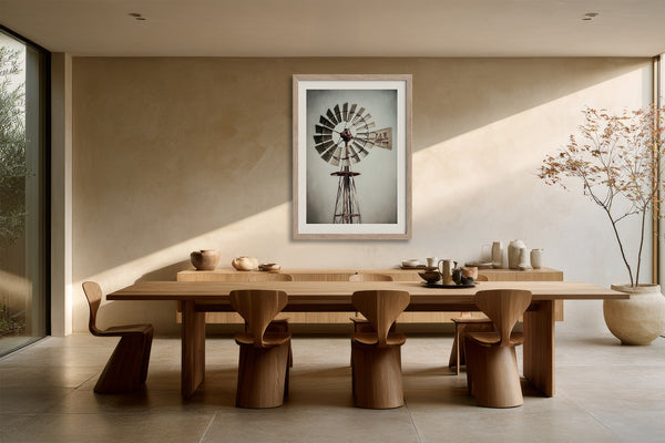 Framed windmill artwork centered above a long wood dining table and sculptural chairs.