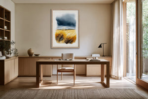Light wood-framed coastal watercolour above a wooden desk, showing golden dune grasses beneath dark storm clouds.