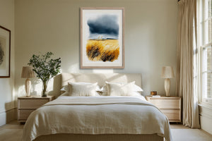 Light wood-framed coastal watercolour of golden grasses and dark clouds displayed above a bed.