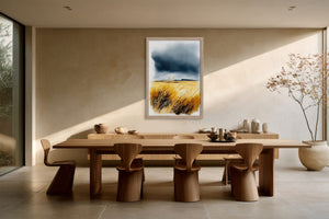 Framed watercolour of golden dune grasses beneath storm clouds hanging above a long wooden dining table.